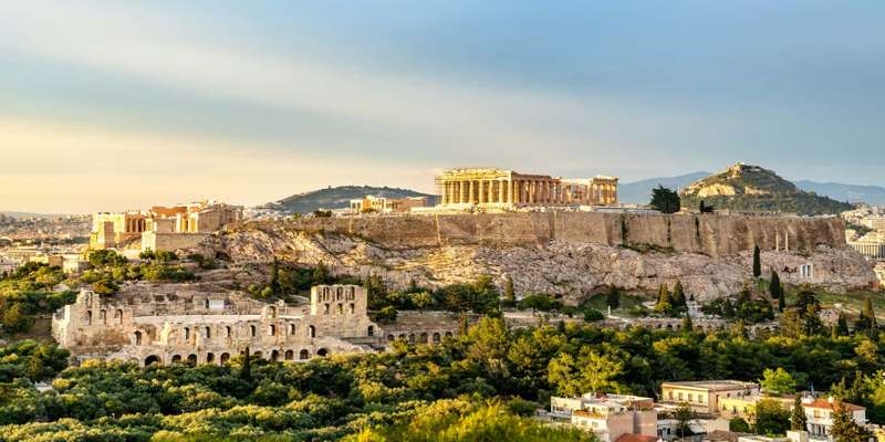 View of the Acropolis and Parthenon rising above Athens cityscape at golden hour with Aegean Sea visible in background, classical heritage travel photography