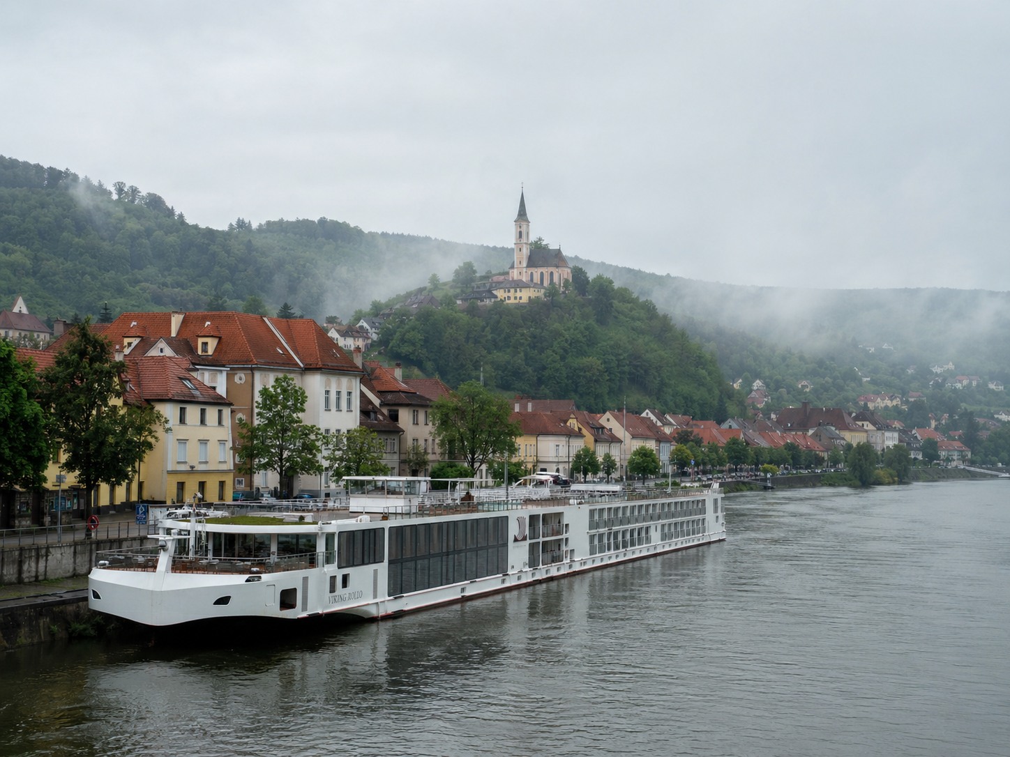 Luxury river cruise ship docked at a small European river town with church spire and forested hills in soft morning mist, Danube embarkation photography