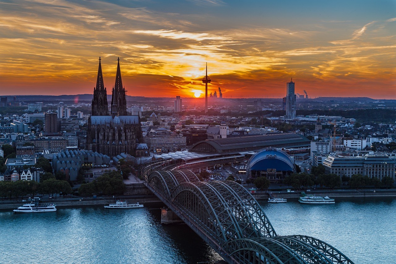Cologne Cathedral twin Gothic spires rising above the Rhine river at sunset with Hohenzollern Bridge and river cruise ships in foreground, German heritage photography