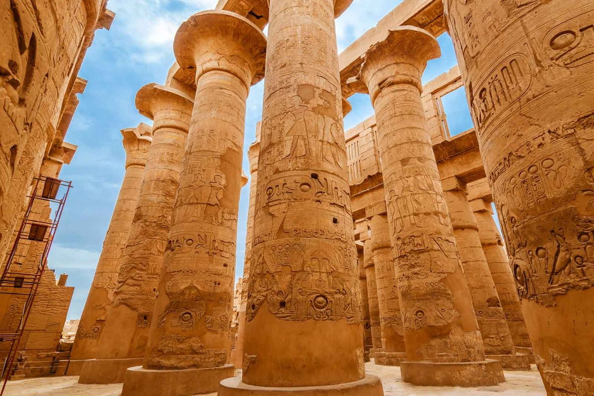 Karnak Temple complex with massive stone columns and hieroglyphic carvings in warm golden afternoon light with desert sand and blue sky, Egyptian archaeological photography