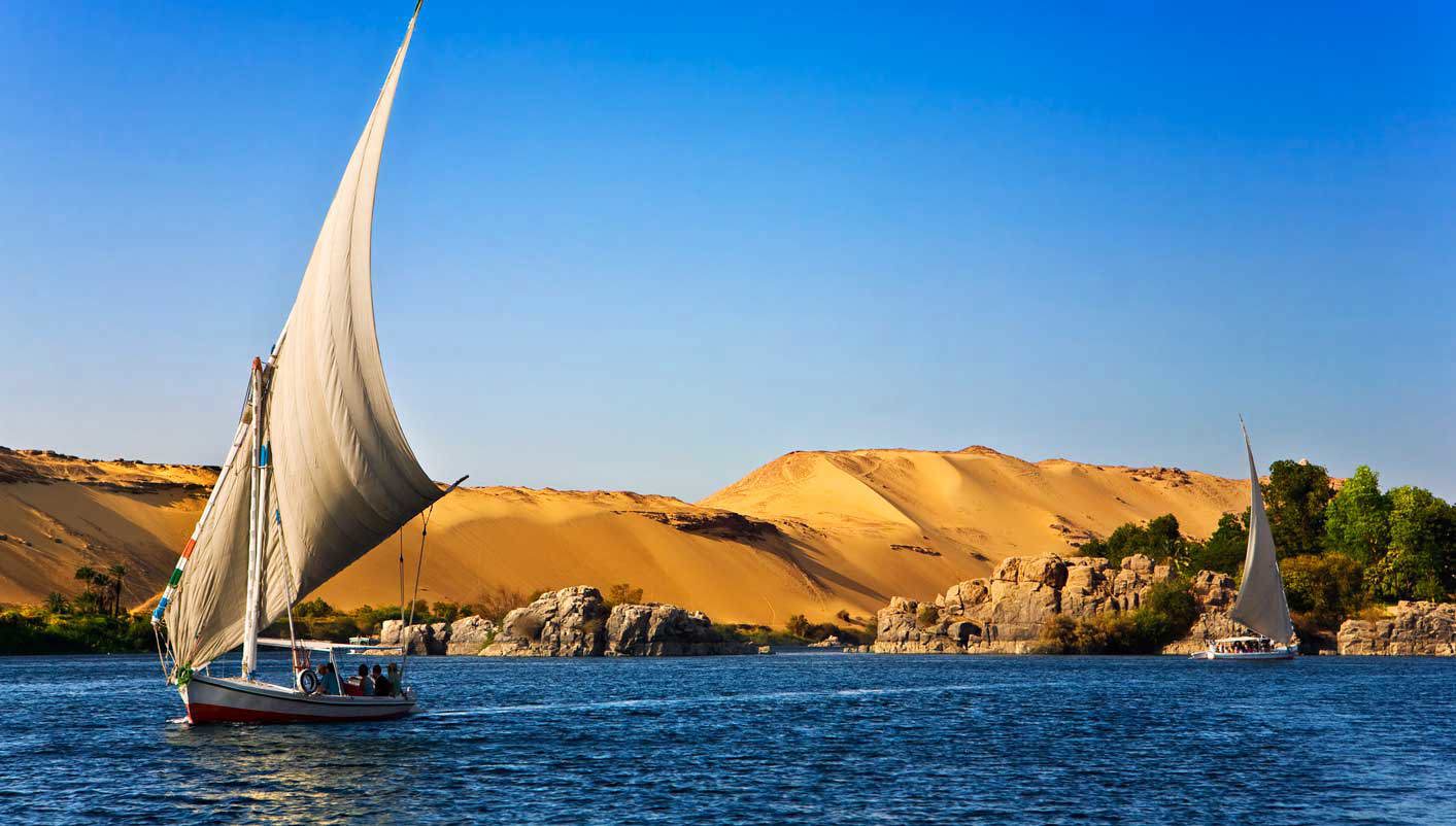 Traditional wooden felucca sailboat on the Nile river at Aswan with golden sand dunes and Aga Khan Mausoleum on the west bank in warm afternoon light, Egyptian sailing photography