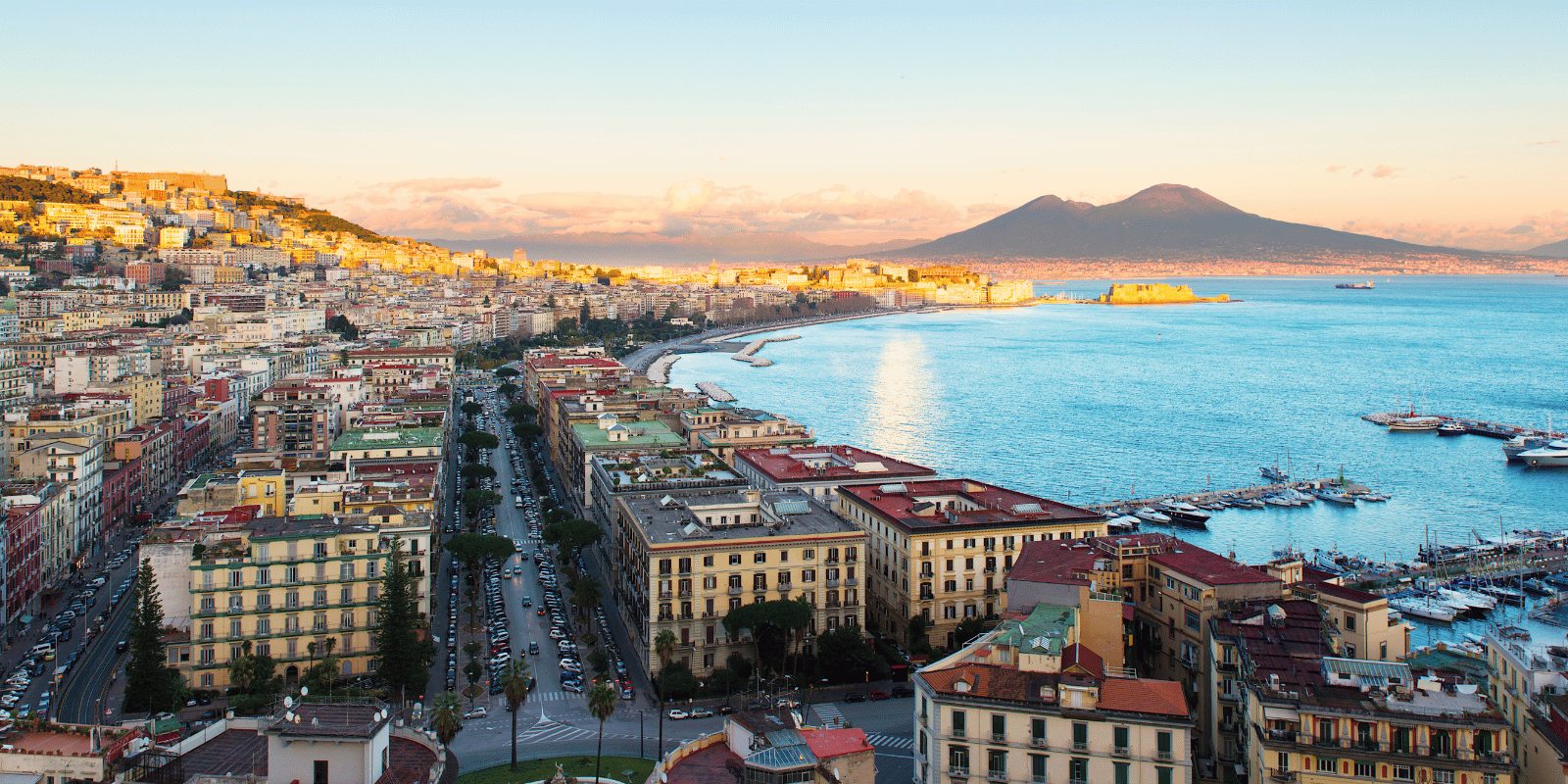 View across the Bay of Naples with Vesuvius rising behind pastel-colored waterfront buildings in warm late afternoon light, Italian coastal photography