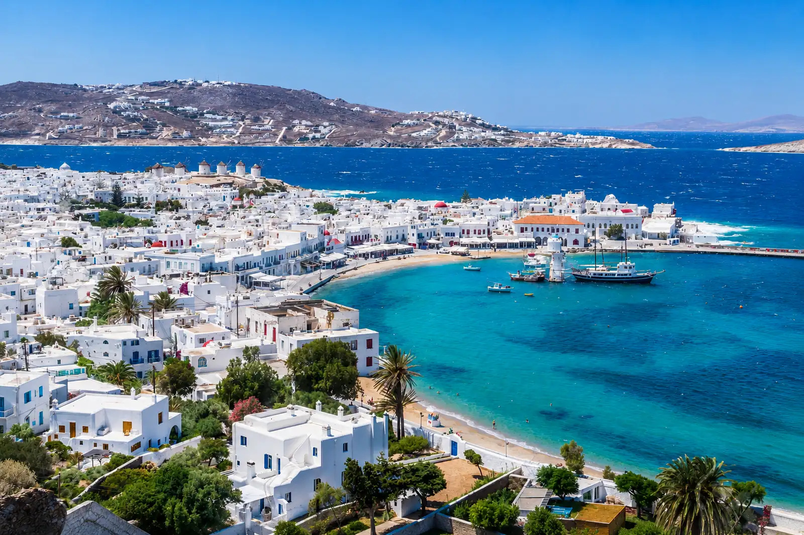 Aerial view of whitewashed Cycladic village on cliff edge overlooking deep blue Aegean Sea with small yacht anchored in turquoise bay below, Greek islands cruise photography