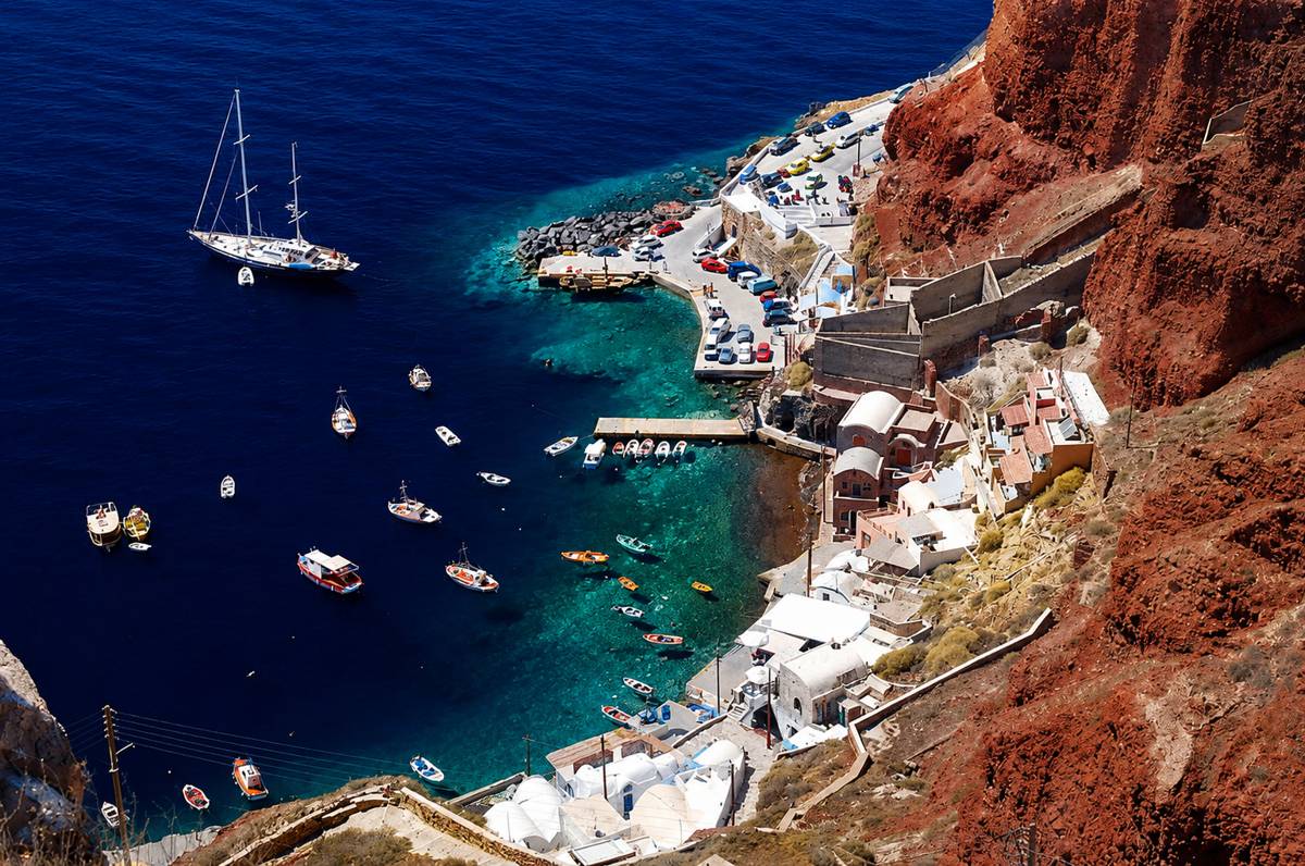 Small cruise yacht entering Santorini caldera at sunset with dramatic volcanic cliffs rising from deep blue water and whitewashed Oia village perched on rim above, Mediterranean travel photography