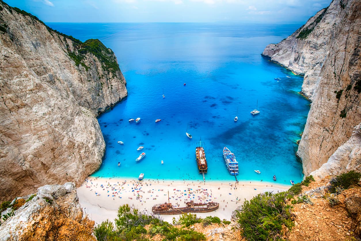 Aerial view of lush green Ionian island coastline with small yacht anchored in sheltered turquoise cove surrounded by cypress trees and limestone cliffs, Greece cruise photography
