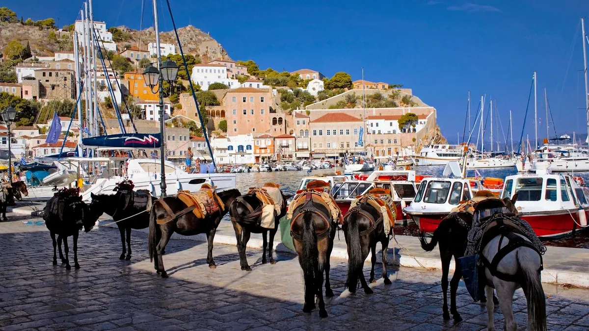 Hydra island harbor with 18th-century stone mansions lining the waterfront and donkeys waiting at the dock, no cars visible, warm afternoon light reflecting on calm water, Greek island photography