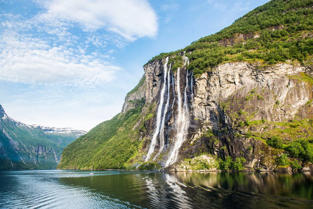 Geirangerfjord aerial view with Seven Sisters waterfall cascading down green cliff face into deep blue fjord water and tiny cruise ship below for scale, UNESCO Norway photography
