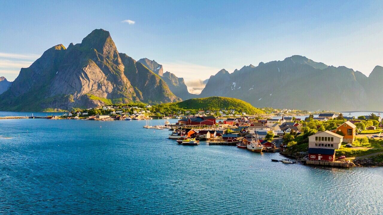 Lofoten islands Reine fishing village with red wooden rorbu cabins at base of dramatic granite peaks reflected in calm fjord water, Arctic summer light, Norwegian travel photography