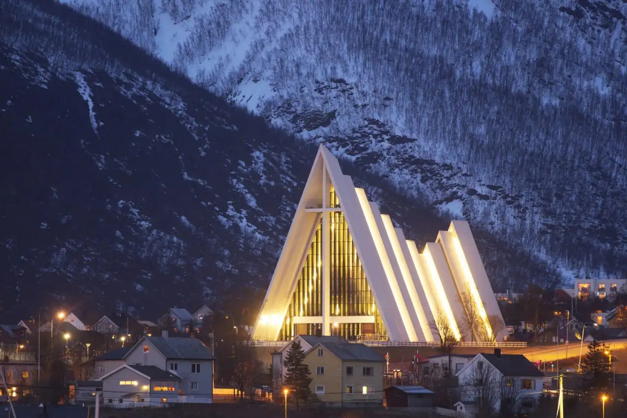 Tromsø Arctic capital cityscape at midnight sun with modern Arctic Cathedral and harbor buildings reflecting in calm water, mountains behind, warm golden Arctic light photography