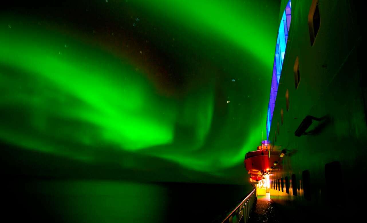 Northern lights aurora borealis green curtain dancing over Norwegian Arctic fjord with snow-covered mountains and expedition ship silhouette reflected in dark water, winter polar photography