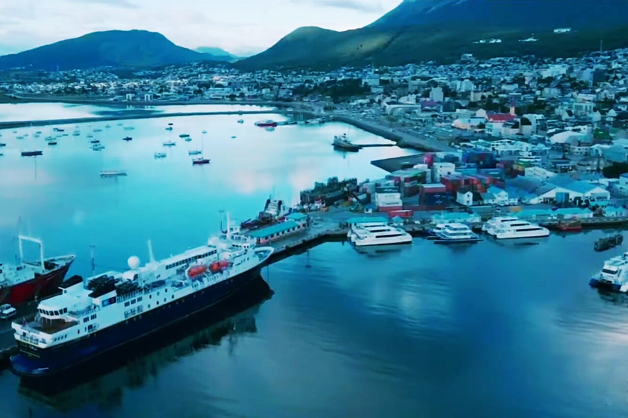 Ushuaia harbor at dusk with expedition ships docked and snow-capped Martial Mountains rising behind the southernmost city, warm lights reflecting on Beagle Channel, travel photography