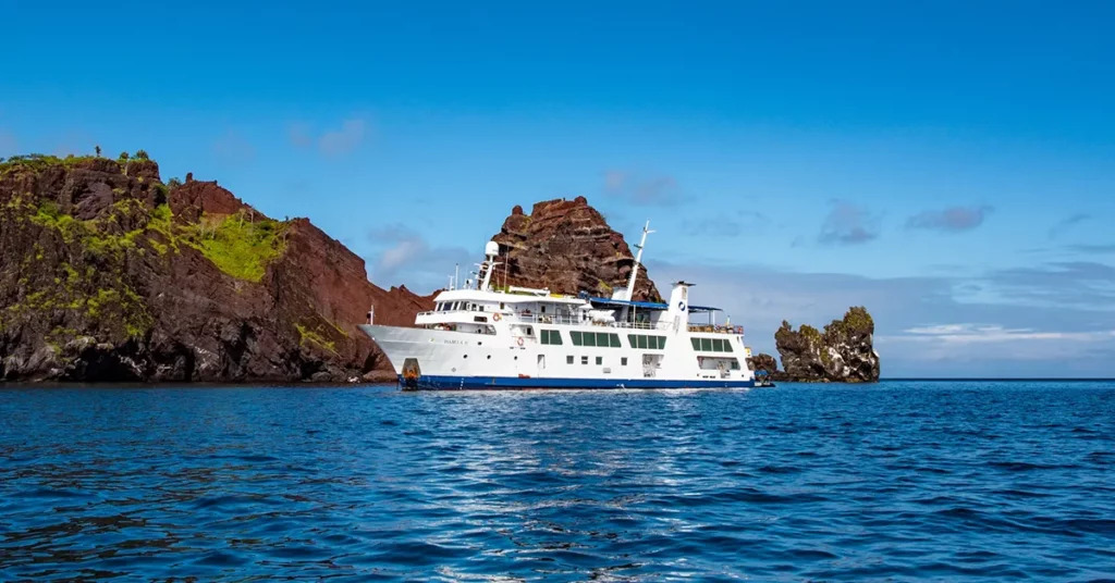 Galápagos Islands aerial view with volcanic coastline meeting deep blue Pacific Ocean and small expedition yacht anchored in turquoise bay, equatorial wildlife sanctuary photography