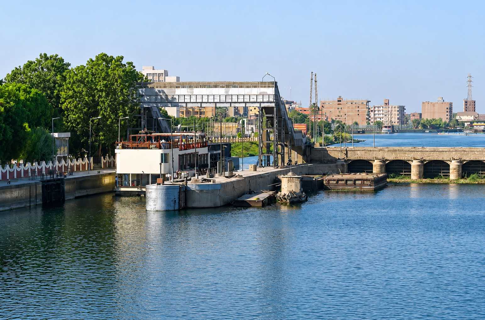 Dahabiya sailboat passing through Esna lock on the Nile River with traditional Egyptian town visible on both banks and local life along the waterfront, authentic river travel photography