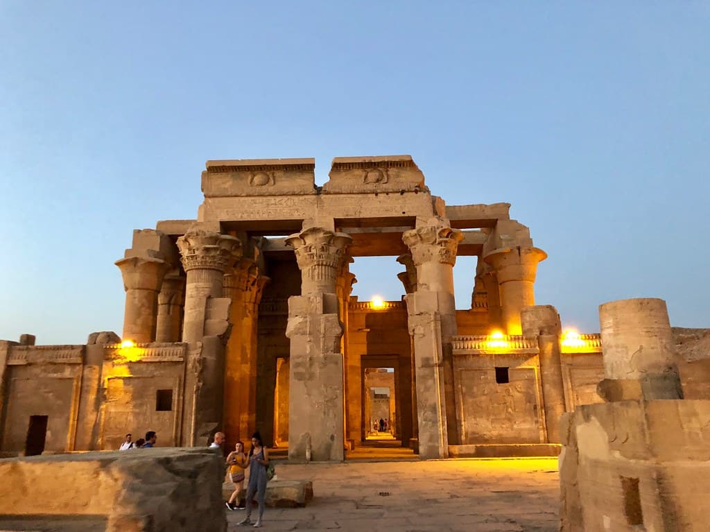 Kom Ombo Temple perched on elevated Nile riverbank at dusk with double entrance columns and crocodile god reliefs illuminated by warm light, dahabiya visible on river below, Egyptian temple photography