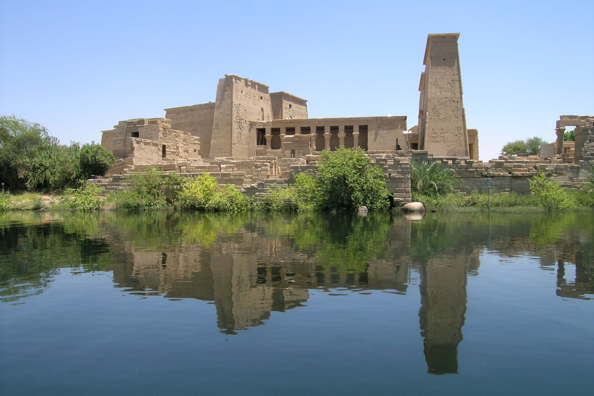 Philae Temple on Agilkia Island near Aswan seen from felucca boat on the Nile with granite boulders and palm trees framing the ancient sanctuary, warm golden hour light, Egyptian heritage photography