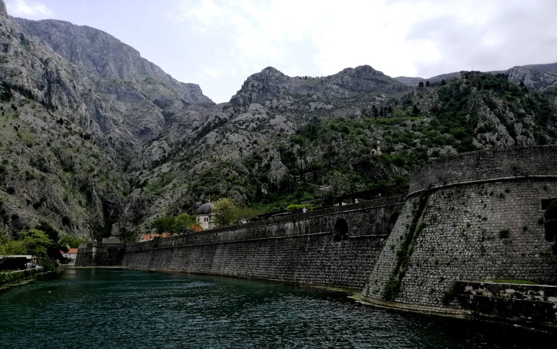 Bay of Kotor fjord-like inlet with medieval fortified town at base of dramatic limestone mountains and calm dark blue water, small ship entering narrow passage, Montenegrin coastal photography