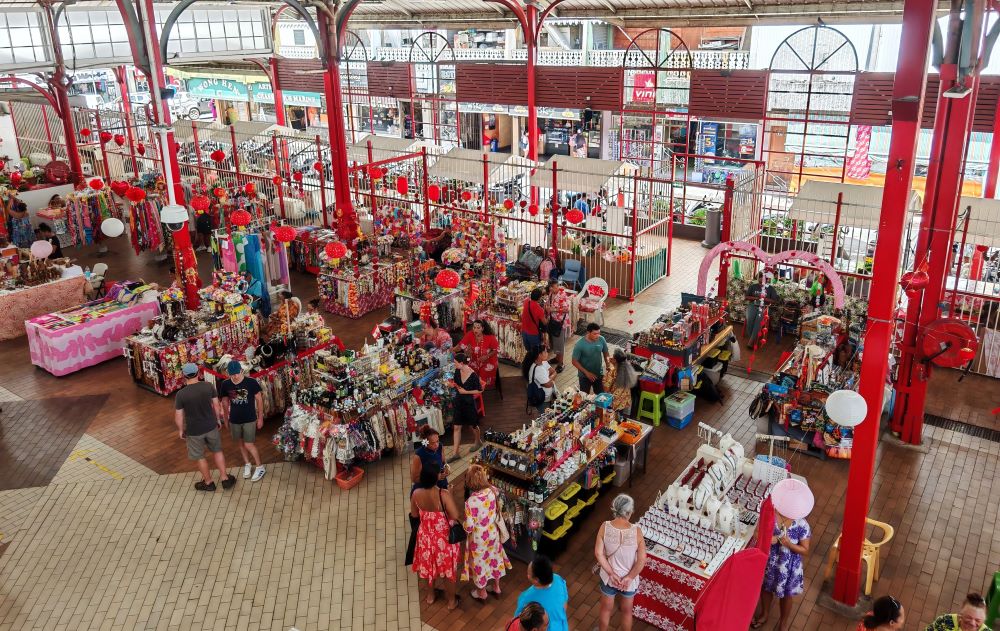 Papeete waterfront market in Tahiti with colorful tropical flowers and fresh fish displayed at stalls, volcanic peaks visible behind the harbor, warm Polynesian morning light, South Pacific travel photography
