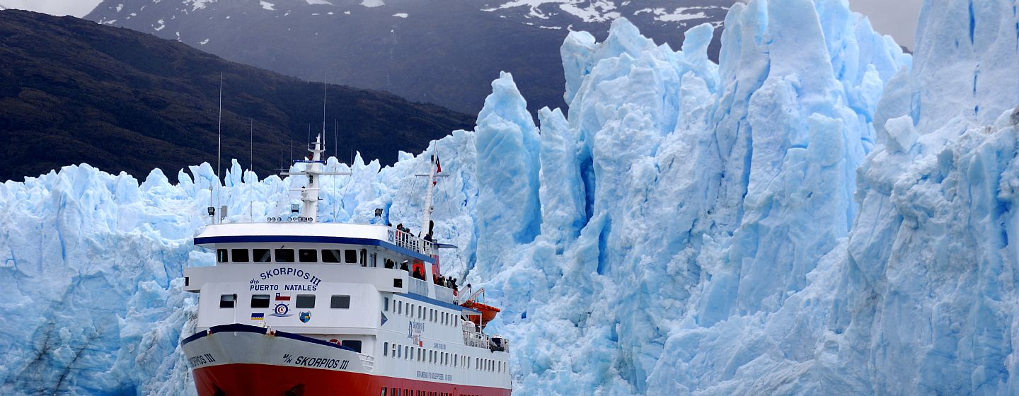 Expedition ship navigating narrow Chilean fjord channel with towering granite walls and hanging glaciers on both sides under dramatic overcast Patagonian sky, South American wilderness cruise photography