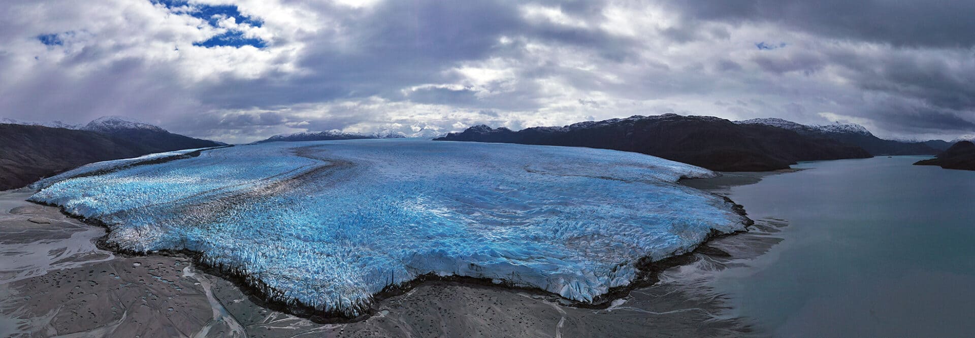 Pio XI Glacier massive blue tidewater face calving ice blocks into Chilean fjord with zodiac expedition boat approaching for scale, mist rising from impact zone, Patagonian glacier expedition photography