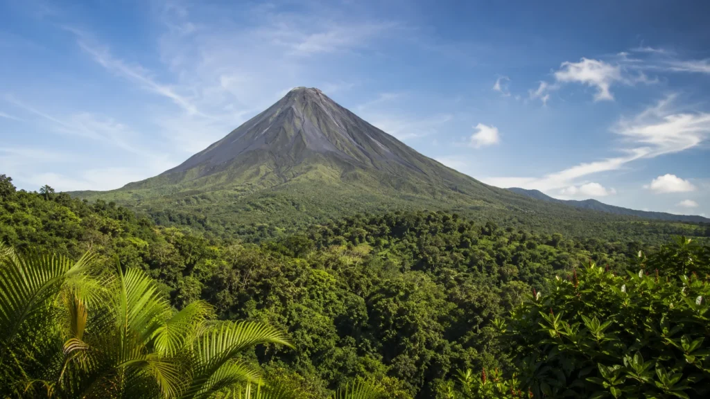 Arenal Volcano Costa Rica rising above lush green rainforest canopy with blue sky and wispy clouds, dramatic landscape advent