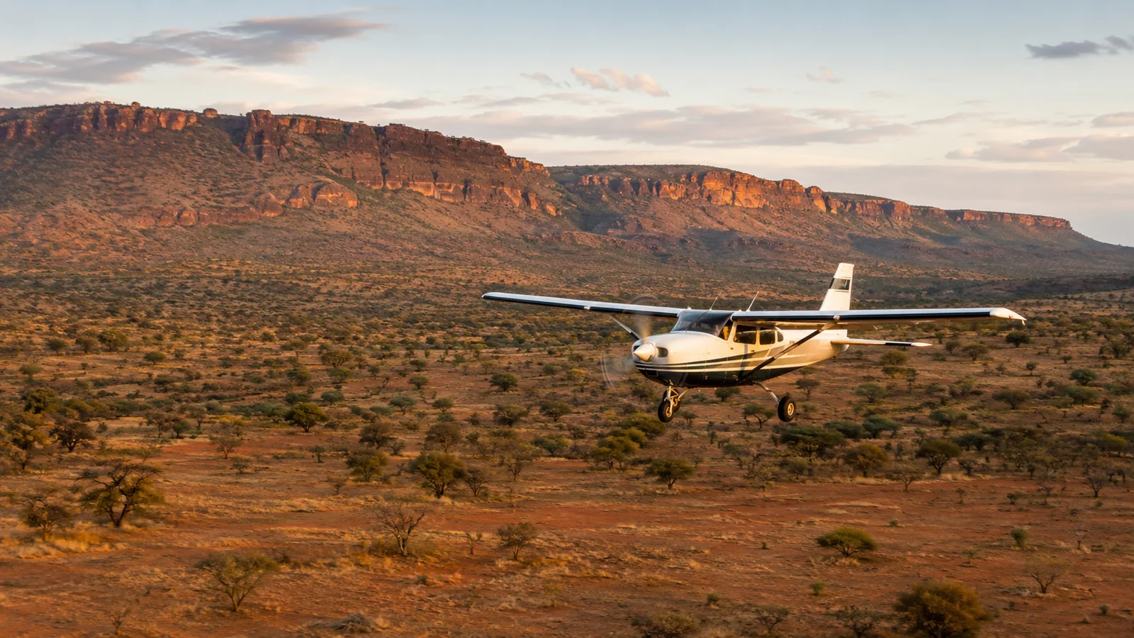 Light aircraft flying low over the red-earth landscape of northern Kenya with scattered acacia trees and the Laikipia escarpment behind, safari transfer travel photography