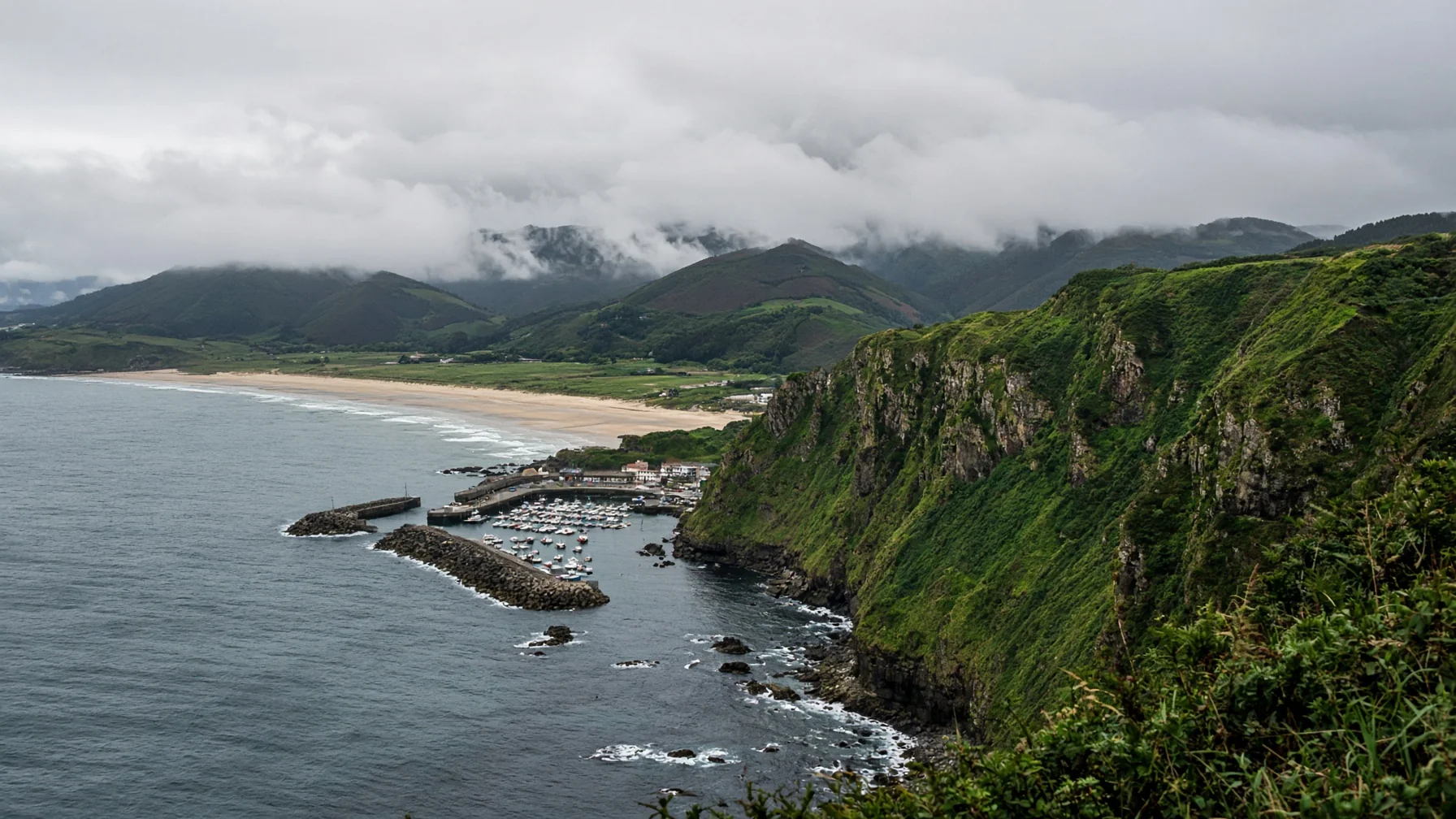 Green cliffs falling to a wide Atlantic beach with low cloud over rolling hills and a small fishing harbour below, editorial travel photography