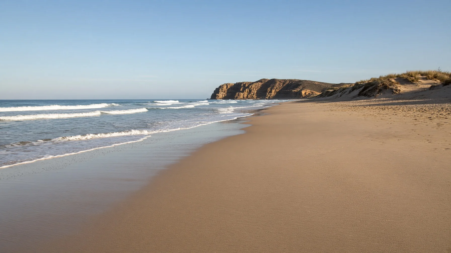 Long empty Atlantic beach with breaking waves, low dunes and a headland in the distance under clear autumn light, editorial travel photography