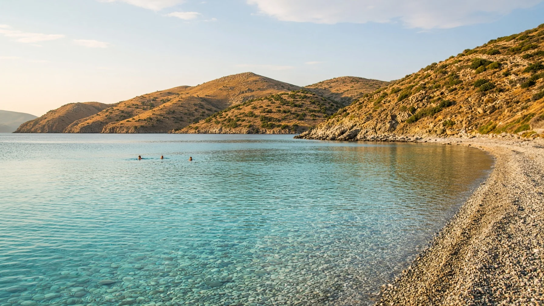 Calm turquoise bay in early autumn with swimmers in clear water, empty pebble shore and warm low sunlight on dry hills behind, serene coastal photography
