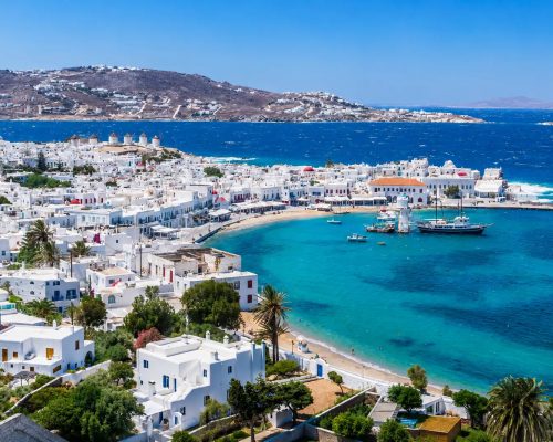 Aerial view of whitewashed Cycladic village on cliff edge overlooking deep blue Aegean Sea with small yacht anchored in turqu