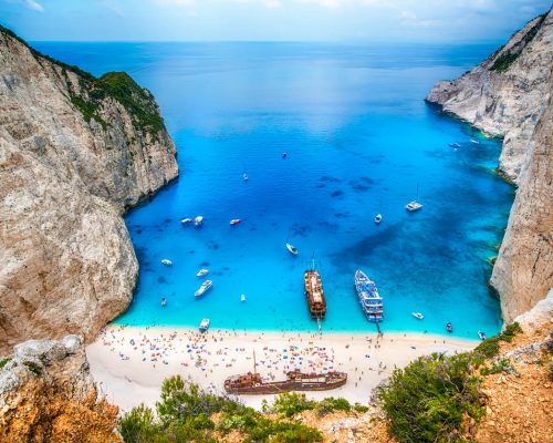 Aerial view of lush green Ionian island coastline with small yacht anchored in sheltered turquoise cove surrounded by cypress