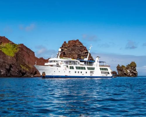 Galápagos Islands aerial view with volcanic coastline meeting deep blue Pacific Ocean and small expedition yacht anchored in
