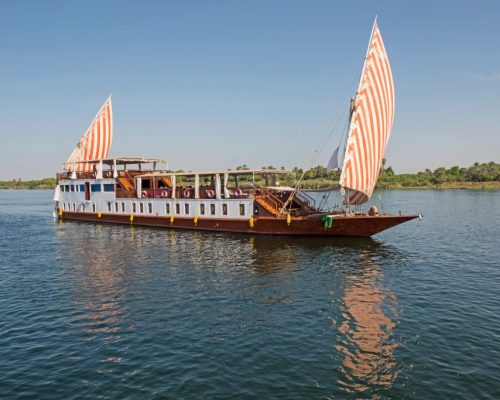Traditional dahabiya sailboat with white sails unfurled cruising the Nile River at sunset with palm trees and ancient temple