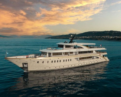 Small cruise ship sailing along dramatic Dalmatian coastline with ancient walled city on rocky peninsula and turquoise Adriat