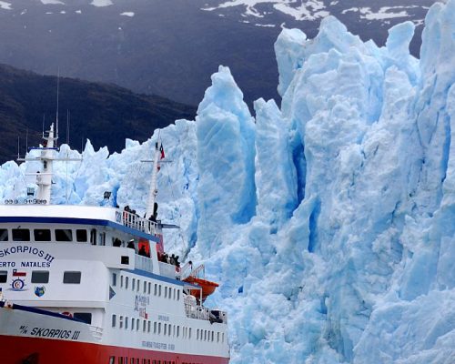Expedition ship navigating narrow Chilean fjord channel with towering granite walls and hanging glaciers on both sides under