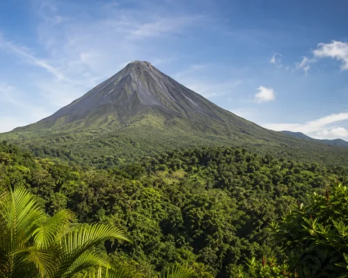 Arenal Volcano Costa Rica rising above lush green rainforest canopy with blue sky and wispy clouds, dramatic landscape advent