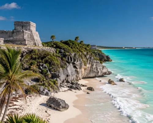 Tulum ruins Maya clifftop temple overlooking turquoise Caribbean Sea with white sand beach and palm trees, bright sky, iconic